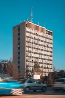 A high-rise office building with a grid of windows and air conditioning units mounted on each floor. Several antennas and communication equipment are installed on the rooftop. The building is surrounded by trees, and there is a billboard in front of it. Cars are moving along the road in the foreground, captured with a sense of motion blur.