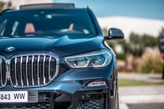 A close-up view of a luxury SUV, showcasing its sleek front design. The image highlights a shiny black exterior with an intricate grille, striking headlights, and a visible license plate. The background is blurred, indicating an outdoor setting with greenery.