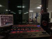 A sound mixing console with colorful backlit buttons and sliders is positioned in front of a glass window, offering a view into a large room where a group of people is gathered, possibly for a performance or event. To the left, a computer monitor displays audio control software, while speakers and other audio equipment are visible around the console.