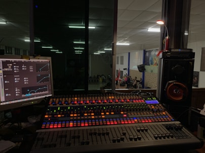 A sound mixing console with colorful backlit buttons and sliders is positioned in front of a glass window, offering a view into a large room where a group of people is gathered, possibly for a performance or event. To the left, a computer monitor displays audio control software, while speakers and other audio equipment are visible around the console.