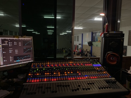 A sound mixing console with colorful backlit buttons and sliders is positioned in front of a glass window, offering a view into a large room where a group of people is gathered, possibly for a performance or event. To the left, a computer monitor displays audio control software, while speakers and other audio equipment are visible around the console.