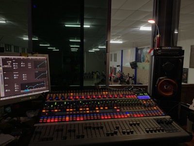 A sound mixing console with colorful backlit buttons and sliders is positioned in front of a glass window, offering a view into a large room where a group of people is gathered, possibly for a performance or event. To the left, a computer monitor displays audio control software, while speakers and other audio equipment are visible around the console.