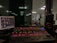 A sound mixing console with colorful backlit buttons and sliders is positioned in front of a glass window, offering a view into a large room where a group of people is gathered, possibly for a performance or event. To the left, a computer monitor displays audio control software, while speakers and other audio equipment are visible around the console.