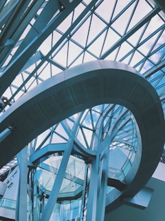 A modern architectural structure featuring a glass and metal atrium with geometric patterns. The curved metallic frame and glass walls create a dynamic and futuristic impression, while the ceiling is composed of large glass panels allowing natural light to enter.