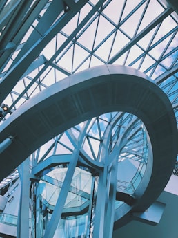 A modern architectural structure featuring a glass and metal atrium with geometric patterns. The curved metallic frame and glass walls create a dynamic and futuristic impression, while the ceiling is composed of large glass panels allowing natural light to enter.