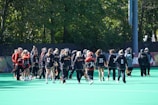A group of athletes in sports uniforms gathers on a field surrounded by trees. They are holding lacrosse sticks and wearing different numbered jerseys, some in black and others in red. Most have their hair tied back, and a few wear visors or headbands. The setting is a bright, sunny day.