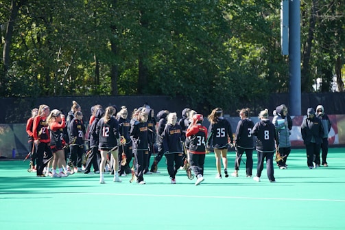 A group of athletes in sports uniforms gathers on a field surrounded by trees. They are holding lacrosse sticks and wearing different numbered jerseys, some in black and others in red. Most have their hair tied back, and a few wear visors or headbands. The setting is a bright, sunny day.