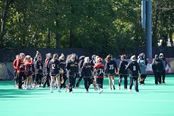 A group of athletes in sports uniforms gathers on a field surrounded by trees. They are holding lacrosse sticks and wearing different numbered jerseys, some in black and others in red. Most have their hair tied back, and a few wear visors or headbands. The setting is a bright, sunny day.