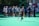 A sports team gathers on a field with a coach, all dressed in athletic gear, while one player wearing a red helmet and jersey walks away from the group carrying a lacrosse stick. The field is green, and there are sports equipment and water bottles visible.