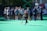 A sports team gathers on a field with a coach, all dressed in athletic gear, while one player wearing a red helmet and jersey walks away from the group carrying a lacrosse stick. The field is green, and there are sports equipment and water bottles visible.