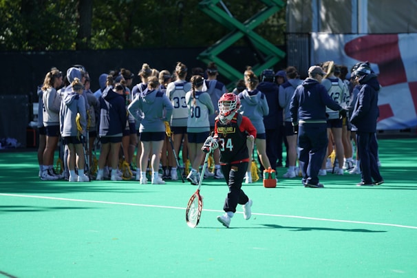 A sports team gathers on a field with a coach, all dressed in athletic gear, while one player wearing a red helmet and jersey walks away from the group carrying a lacrosse stick. The field is green, and there are sports equipment and water bottles visible.
