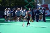 A sports team gathers on a field with a coach, all dressed in athletic gear, while one player wearing a red helmet and jersey walks away from the group carrying a lacrosse stick. The field is green, and there are sports equipment and water bottles visible.