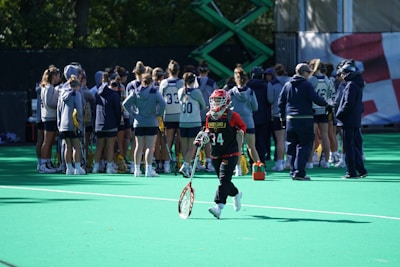 A sports team gathers on a field with a coach, all dressed in athletic gear, while one player wearing a red helmet and jersey walks away from the group carrying a lacrosse stick. The field is green, and there are sports equipment and water bottles visible.