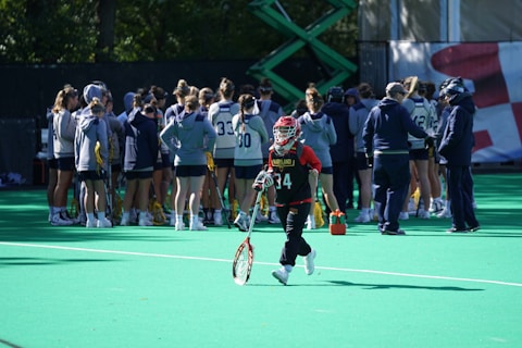 A sports team gathers on a field with a coach, all dressed in athletic gear, while one player wearing a red helmet and jersey walks away from the group carrying a lacrosse stick. The field is green, and there are sports equipment and water bottles visible.