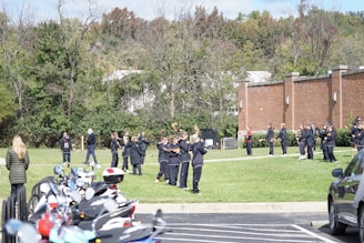 A group of people, likely a sports team, is gathered outdoors on a grassy area. They are dressed in sports attire with some holding lacrosse sticks. The location is beside a brick building, and there are trees in the background. A line of parked motorcycles is visible in the foreground.