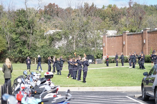 A group of people, likely a sports team, is gathered outdoors on a grassy area. They are dressed in sports attire with some holding lacrosse sticks. The location is beside a brick building, and there are trees in the background. A line of parked motorcycles is visible in the foreground.
