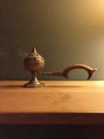 A brass incense burner with a perforated, round lid is placed on a wooden surface. It features an elongated, curved handle made of wood. The background is a plain olive green wall, creating a warm ambiance.