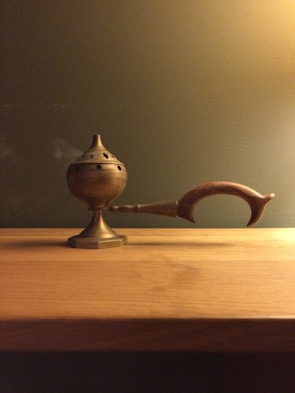 A brass incense burner with a perforated, round lid is placed on a wooden surface. It features an elongated, curved handle made of wood. The background is a plain olive green wall, creating a warm ambiance.
