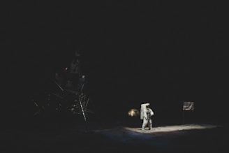 An astronaut stands on a dark lunar surface next to a flag, bathed in the faint illumination of the scene. A lunar module is partially visible in the shadowy background.
