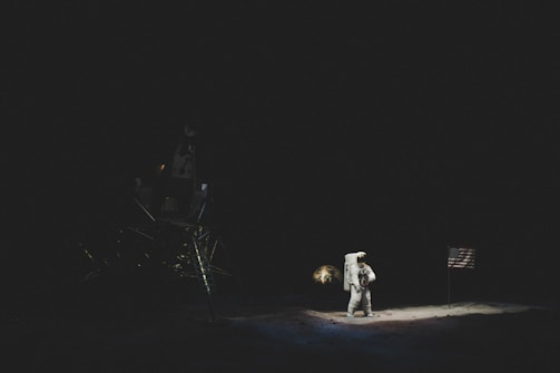 An astronaut stands on a dark lunar surface next to a flag, bathed in the faint illumination of the scene. A lunar module is partially visible in the shadowy background.