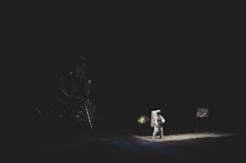 An astronaut stands on a dark lunar surface next to a flag, bathed in the faint illumination of the scene. A lunar module is partially visible in the shadowy background.