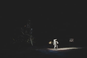 An astronaut stands on a dark lunar surface next to a flag, bathed in the faint illumination of the scene. A lunar module is partially visible in the shadowy background.