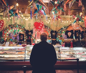 A person stands in front of a colorful bakery display in a festive setting, surrounded by hanging piñatas and vibrant decorations. The display case is filled with a variety of pastries and sweets, illuminated by warm lighting. The background is lively with lights and decorations, featuring heart shapes and various ornamental items.
