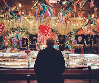 A person stands in front of a colorful bakery display in a festive setting, surrounded by hanging piñatas and vibrant decorations. The display case is filled with a variety of pastries and sweets, illuminated by warm lighting. The background is lively with lights and decorations, featuring heart shapes and various ornamental items.
