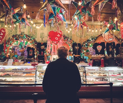 A person stands in front of a colorful bakery display in a festive setting, surrounded by hanging piñatas and vibrant decorations. The display case is filled with a variety of pastries and sweets, illuminated by warm lighting. The background is lively with lights and decorations, featuring heart shapes and various ornamental items.
