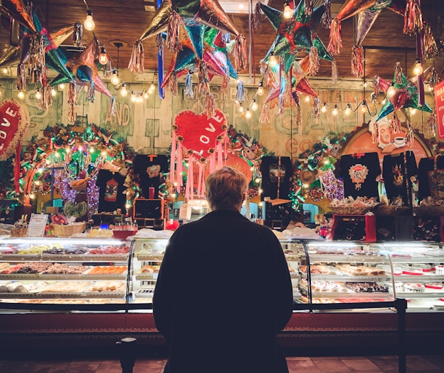 A person stands in front of a colorful bakery display in a festive setting, surrounded by hanging piñatas and vibrant decorations. The display case is filled with a variety of pastries and sweets, illuminated by warm lighting. The background is lively with lights and decorations, featuring heart shapes and various ornamental items.