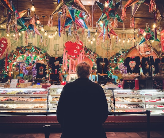 A person stands in front of a colorful bakery display in a festive setting, surrounded by hanging pi&ntilde;atas and vibrant decorations. The display case is filled with a variety of pastries and sweets, illuminated by warm lighting. The background is lively with lights and decorations, featuring heart shapes and various ornamental items.