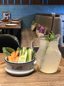 A wooden table displays a bowl filled with fresh vegetable sticks, including carrots and cucumbers, accompanied by a garnish of basil leaves. Beside it is a clear glass pitcher filled with a refreshing lemonade, ice cubes, and a sprig of mint. The background features a cozy restaurant setting with a blue-tiled wall, upholstered seating, and colorful pillows.