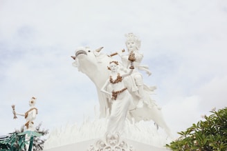 A beautifully detailed white statue features two celestial figures adorned with ornate headdresses and jewelry, sitting on an elaborately carved mythical creature. The scene is set against a soft, cloudy sky and includes lush greenery at the bottom right. The artistry showcases intricate patterns and textures.