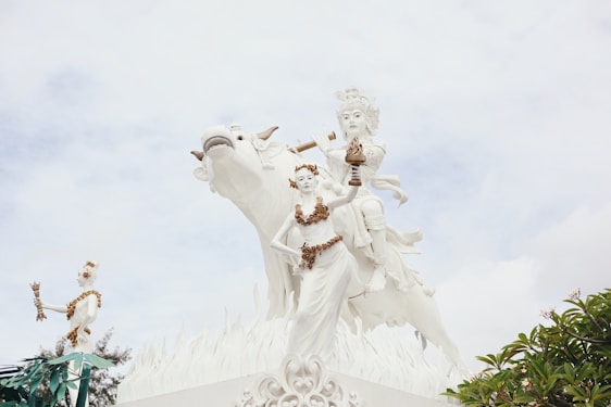 A beautifully detailed white statue features two celestial figures adorned with ornate headdresses and jewelry, sitting on an elaborately carved mythical creature. The scene is set against a soft, cloudy sky and includes lush greenery at the bottom right. The artistry showcases intricate patterns and textures.