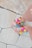 A pair of feet wearing sandals decorated with colorful pom-poms and beads are visible on a tiled pavement. The toes are painted with red nail polish, and the sandals have straps in a neutral color.