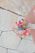 A pair of feet wearing sandals decorated with colorful pom-poms and beads are visible on a tiled pavement. The toes are painted with red nail polish, and the sandals have straps in a neutral color.