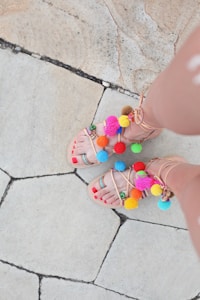A pair of feet wearing sandals decorated with colorful pom-poms and beads are visible on a tiled pavement. The toes are painted with red nail polish, and the sandals have straps in a neutral color.