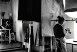 A black and white photograph depicts a studio setting where one person is focused on a camera, possibly adjusting or photographing something on a table. In the background, another person is seated at a desk with a laptop, appearing to be in deep thought or concentration. The room is filled with studio equipment, including light stands and canvases, and has a creative, industrial ambiance.