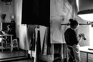 A black and white photograph depicts a studio setting where one person is focused on a camera, possibly adjusting or photographing something on a table. In the background, another person is seated at a desk with a laptop, appearing to be in deep thought or concentration. The room is filled with studio equipment, including light stands and canvases, and has a creative, industrial ambiance.