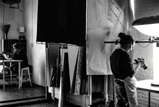 A black and white photograph depicts a studio setting where one person is focused on a camera, possibly adjusting or photographing something on a table. In the background, another person is seated at a desk with a laptop, appearing to be in deep thought or concentration. The room is filled with studio equipment, including light stands and canvases, and has a creative, industrial ambiance.