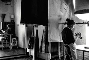 A black and white photograph depicts a studio setting where one person is focused on a camera, possibly adjusting or photographing something on a table. In the background, another person is seated at a desk with a laptop, appearing to be in deep thought or concentration. The room is filled with studio equipment, including light stands and canvases, and has a creative, industrial ambiance.
