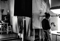 A black and white photograph depicts a studio setting where one person is focused on a camera, possibly adjusting or photographing something on a table. In the background, another person is seated at a desk with a laptop, appearing to be in deep thought or concentration. The room is filled with studio equipment, including light stands and canvases, and has a creative, industrial ambiance.