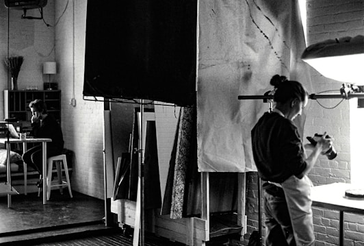 A black and white photograph depicts a studio setting where one person is focused on a camera, possibly adjusting or photographing something on a table. In the background, another person is seated at a desk with a laptop, appearing to be in deep thought or concentration. The room is filled with studio equipment, including light stands and canvases, and has a creative, industrial ambiance.