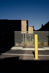 A tall yellow bollard stands in the foreground on a paved street, casting a long shadow. Behind it, a chain-link fence encloses a section of sparse vegetation. Brick buildings with flat roofs are partially illuminated by sunlight, contrasting against the dark shadows. The sky is clear and deep blue.