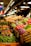 A vibrant market display filled with an array of fresh produce. Colorful vegetables such as tomatoes, carrots, Brussels sprouts, cabbages, and a variety of leafy greens are neatly arranged. Signs with prices are visible, and the scene is well-lit by overhead lamps, creating a welcoming atmosphere.