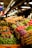 A vibrant market display filled with an array of fresh produce. Colorful vegetables such as tomatoes, carrots, Brussels sprouts, cabbages, and a variety of leafy greens are neatly arranged. Signs with prices are visible, and the scene is well-lit by overhead lamps, creating a welcoming atmosphere.