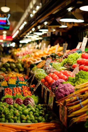 A vibrant market display filled with an array of fresh produce. Colorful vegetables such as tomatoes, carrots, Brussels sprouts, cabbages, and a variety of leafy greens are neatly arranged. Signs with prices are visible, and the scene is well-lit by overhead lamps, creating a welcoming atmosphere.