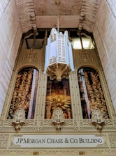 An ornate, intricately designed building entrance features elaborate golden architectural details with decorative carvings on the facade. A large and lavish floral chandelier hangs above the entrance. Below the architectural embellishments is a sign that reads 'JP Morgan Chase & Co. Building'. The entrance is framed by stone walls that add to the grandeur and historic appearance of the site.