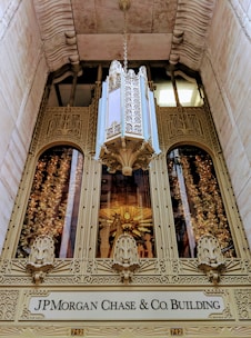 An ornate, intricately designed building entrance features elaborate golden architectural details with decorative carvings on the facade. A large and lavish floral chandelier hangs above the entrance. Below the architectural embellishments is a sign that reads 'JP Morgan Chase & Co. Building'. The entrance is framed by stone walls that add to the grandeur and historic appearance of the site.