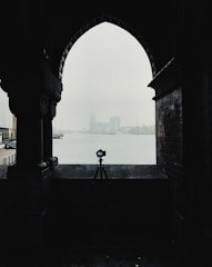 A silhouette of a camera on a tripod is framed by an archway, with a misty waterfront and distant buildings in the background. The scene is moody and atmospheric, with minimal visibility due to the fog.